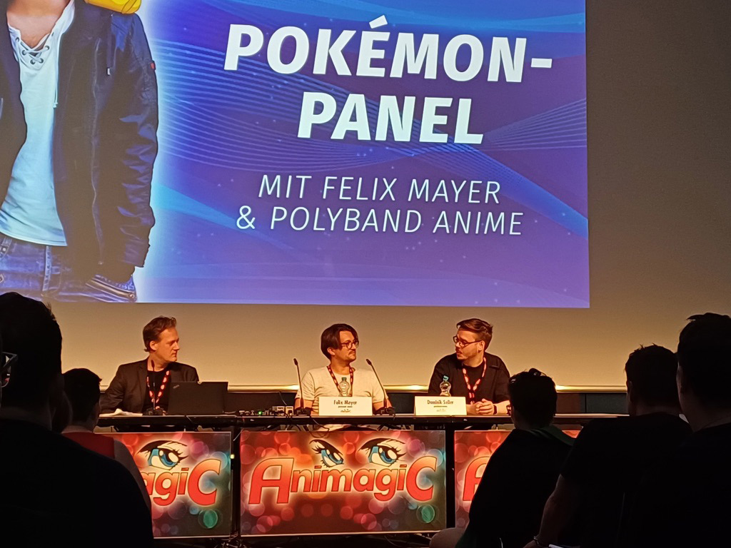 Dominik Seiler moderiert das Pokémon-Panel auf der AnimagiC 2024. Auf der Bühne sitzen Felix Mayer, die deutsche Stimme von Ash Ketchum, und Matthias Süß von Polyband. Alle drei sind in ein Gespräch vertieft.